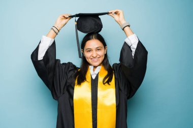 Gülümseyen, heyecanlı Latin kadın mezuniyet kepi ve cübbesini takıp üniversite diplomasını almak için törene hazırlanıyor.