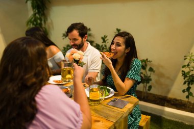 Neşeli mutlu Latin kadınlar ve erkek arkadaşlar eğlenceli bir kutlama ve arka bahçede akşam yemeği sırasında lezzetli pizzalar yiyorlar.