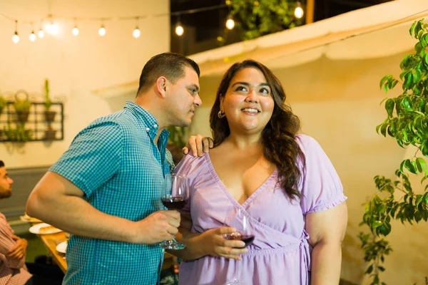Hispanic man whispering a secret to a young woman drinking wine and flirting during a celebration at night with friends