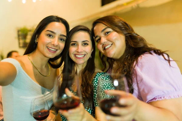 Attractive young women smiling while taking a selfie together while having fun drinking wine at night during a social gathering