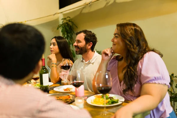 Group of happy friends sitting at the table while eating dinner and drinking wine while talking and celebrating