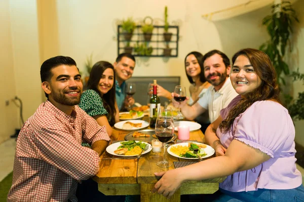 Happy smiling group of six friends sitting at the table ready to eat dinner and drink wine having fun during a celebration