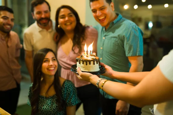 Friends giving a surprise party to a happy hispanic woman holding a birthday cake with candles while singing happy birthday