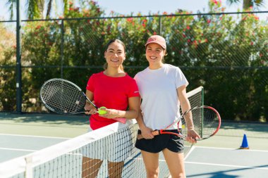 Birlikte tenis oynadıktan sonra raket kullanırken mutlu bir şekilde kameraya bakan heyecanlı genç kadınlar.