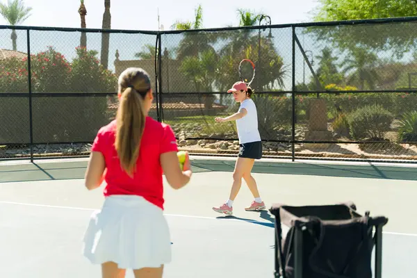 Female tennis coach seen from behind teaching a lesson to a caucasian young woman learning to play tennis
