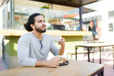 Rahatlamış İspanyol genç adam gülümsüyor açık kafede kahve içerken rahatlıyor ve tatlı yiyor.