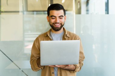 Bir Latin ofis çalışanının portresi ayakta dururken bir dizüstü bilgisayar kullanmaktadır.