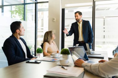 İş adamları, takım elbiseli bir adamı toplantı odasında iş teklifini paylaşırken dinliyor.