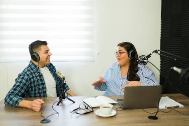 Mutlu kadın ve erkek radyo sunucuları podcast yaparken gülümsüyor ve stüdyoda eğlenceli bir hikayeden bahsediyorlar.
