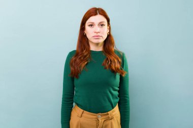 30 'lu yaşlarındaki kızıl saçlı ciddi bir kadın mavi bir stüdyonun arka planında günlük giysiler giymiş kameraya bakıyor.