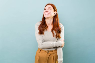 Neşeli kızıl saçlı beyaz kadın heyecanlı görünüyor ve mavi bir stüdyonun önünde eğleniyor.