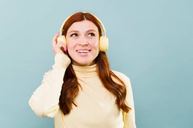Neşeli kızıl saçlı kadın gülümsüyor ve mavi arka planda kulaklıkla müzik dinleyerek eğleniyor.