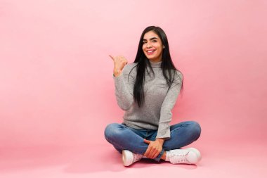 Mutlu ve çekici genç bir kadın yerde oturmuş gülümseyen ve rahatlatan pembe bir fotokopi alanını işaret ediyor. 