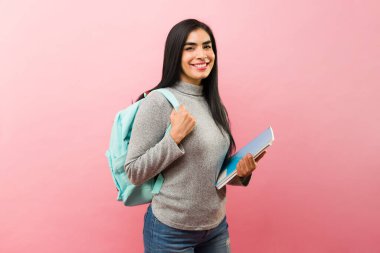 Üniversitede okurken gülümseyen İspanyol üniversite öğrencisi pembe bir arka planda kitap ve sırt çantası taşıyor.