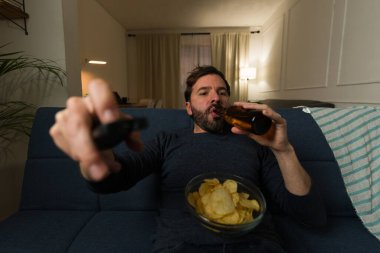 30 'lu yaşlarda rahat bir Latin adam uzaktan kumandayla televizyon izleyip cips yiyip kanepede bira içiyor.