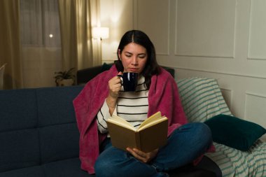 Güzel, beyaz bir kadın kanepeye sarılı sıcak çay içerken rahatlatıcı bir kitap okuyor.