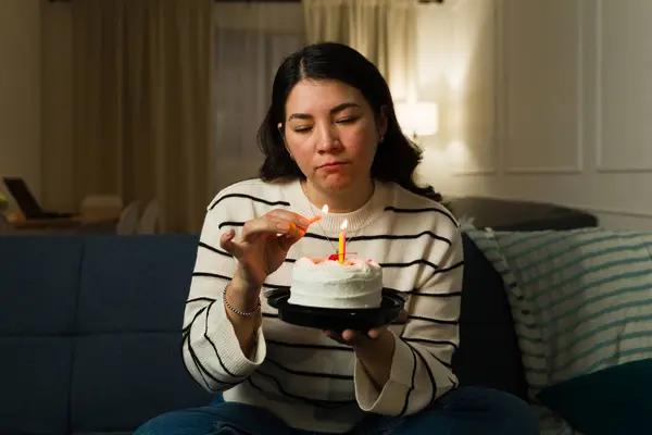 Sad lonely caucasian woman celebrating her birthday alone at home while lighting candles on a cake feeling depressed