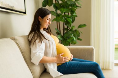 Güzel, hamile karnına bakıp oturma odasında kanepede otururken gülümseyen bir anne bekliyor.