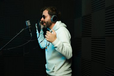 Kulaklık takan neşeli erkek şarkıcı, ses geçirmez bir stüdyoda mikrofonda şarkı söylüyor.