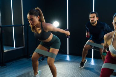 Çağdaş bir jimnastik salonunda insanlar yoğun bir fitness dersi sırasında direniş bantlarıyla çömelme hareketleri yapıyorlar.