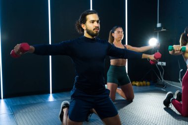 İnsanlar kapalı bir spor salonunda ağırlıklarla çömelme hareketleri yapıyorlar.