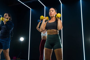 Modern bir spor salonunda dambıllarla çalışan bir grup odaklanmış insan.