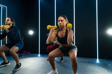 Odaklanmış bireyler, enerjik fitness sınıfında dambıllarla çömelme hareketleri yaparlar.