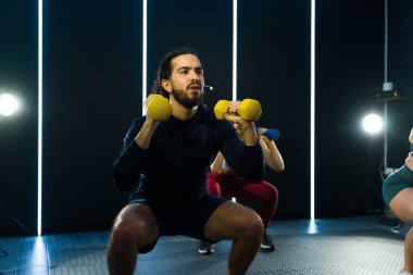Bireyler dinamik fitness sınıfına katılıyorlar, ağırlıklarla çömelme hareketleri yapıyorlar.