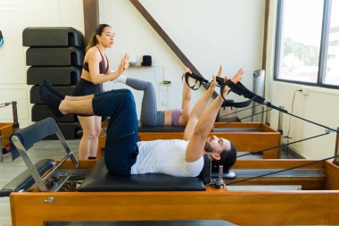 İki birey pilates reformatör yatağında egzersiz yapıyor. Güç ve konsantrasyon gösteriyor.