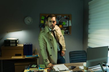 Trençkotlu kendinden emin özel dedektif loş bir ofiste duruyor ve etrafındaki kanıtları inceliyor.