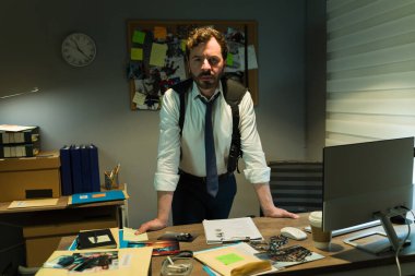 Odaklanmış müfettiş masasının arkasında duruyor. Dava dosyaları ve ipuçlarıyla çevrili. Kararlılığı somutlaştırıyor.