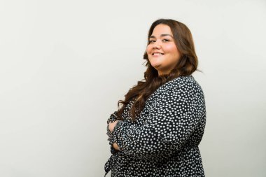 Gülümseyen, kollarını kavuşturup kollarını kavuşturmuş, tarafsız bir arkaplana yaslamış neşeli, iri yarı bir kadın.