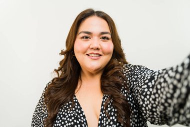 Büyük beden bir kadın beyaz bir arka planda özçekim çekerken kendine güven ve neşe saçıyor.