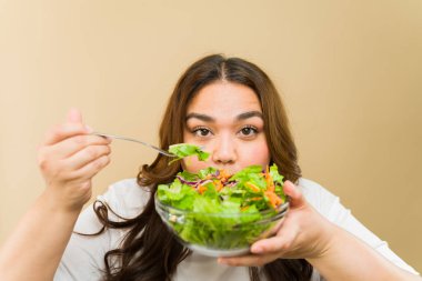 Kıvrımlı vücutlu mutlu genç bir kadın basit bir arka planda besleyici bir salatanın tadını çıkarıyor.