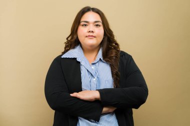 İş elbisesi giymiş, bronz bir arka planda çapraz kollarla poz veren iddialı, iri yarı bir kadının portresi.