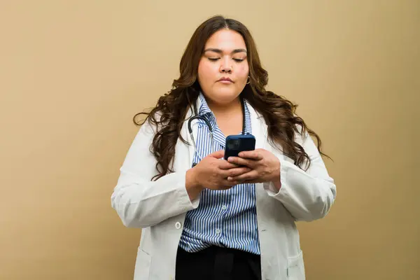 Confident plus-size female doctor in lab coat captivates attention with her phone while exuding style