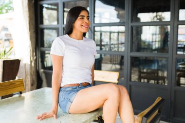 Basit beyaz tişörtlü mutlu Latin kadın. Model modeller için ideal. Kafe masasında eğleniyor, uzaklığa bakıyor.
