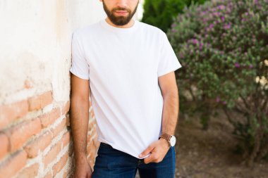 Modaya uygun İspanyol adam, kentin tuğla duvarına karşı markalaştırmaya hazır sade beyaz bir tişört sunuyor.