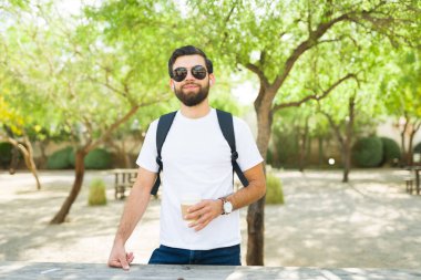 Sırt çantasıyla kendine güveni tam genç İspanyol bir adam, modellik için beyaz tişört giyiyor, güneş gözlüğü takıyor ve güneşli bir park ortamında gülümsüyor.