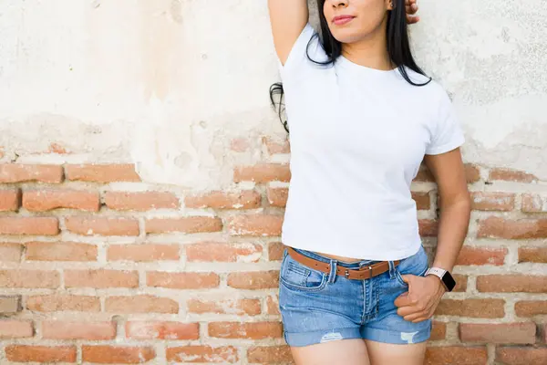 白い白いTシャツを着ているラテン系女性のクローズアップは デザインのモックアップやファッションプレゼンテーションに最適な素朴なレンガ壁に立ちます ロイヤリティフリーのストック画像