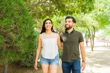 Mutlu, genç, İspanyol bir çift birlikte yürüyor, gülümsüyor ve el ele tutuşuyorlar, güneşli bir günde, açık havada.