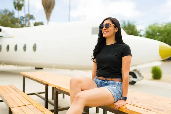 Self-assured latina woman in a black t-shirt and sunglasses sitting casually in front of a small airplane on a sunny day