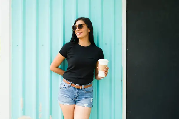 コーヒーカップを保持する黒いTシャツでヒスパニック女性を笑うことは 晴れた日にサングラスをかけたティールの背景に自信を持って立っています ロイヤリティフリーのストック画像