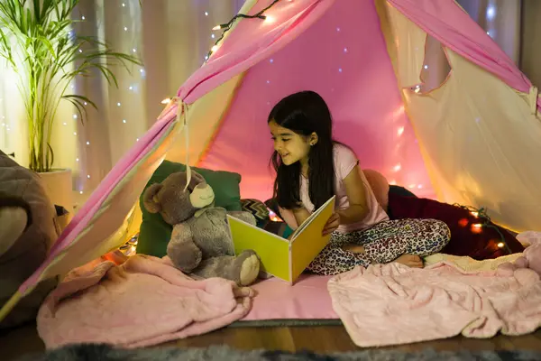 リトルヒスパニックの少女は 夜の寝室で自家製のテントで彼女のテディベアに本を読むのが好きです ストック写真