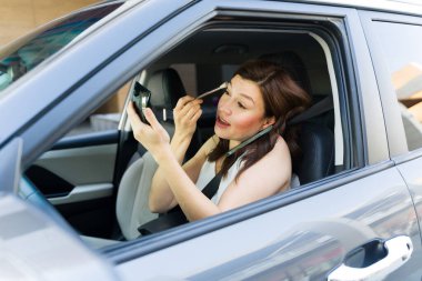 Odaklanmış kadın çoklu görevleri, arabadaki fırçayla makyaj yapmak, modern hayatın koşuşturma ve güvenli sürüş normlarına örnek teşkil etmek.