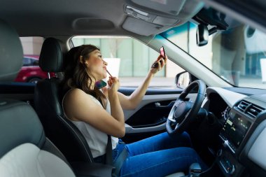 Odaklanmış genç bir kadın elinde telefon tutarken ruj sürüyor. Araba kullanırken birden fazla iş yapmanın riskli davranışlarını gösteriyor.