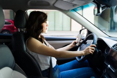 Beyaz bir kadın bir elinde akıllı bir telefonla direksiyonda dikkatle araba kullanıyor.