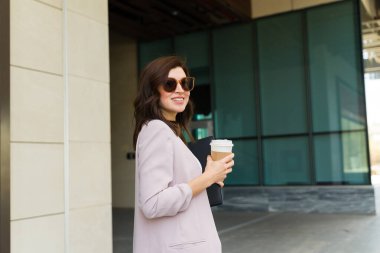 Güneş gözlüklü profesyonel bir kadın dışarıda kahve yudumluyor, şehir yaşamını somutlaştırıyor ve şehir içi seyahatlerin dinamik doğası