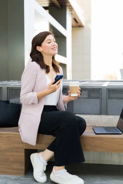 Relaxed Professional Businesswoman Smiles While Holding Her Smartphone Coffee Seated รูปภาพสต็อก