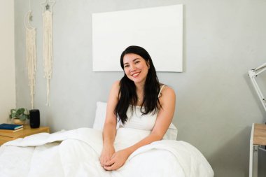 Joyful woman with a captivating smile sits comfortably on her bed, exuding relaxation and happiness in a serene bedroom